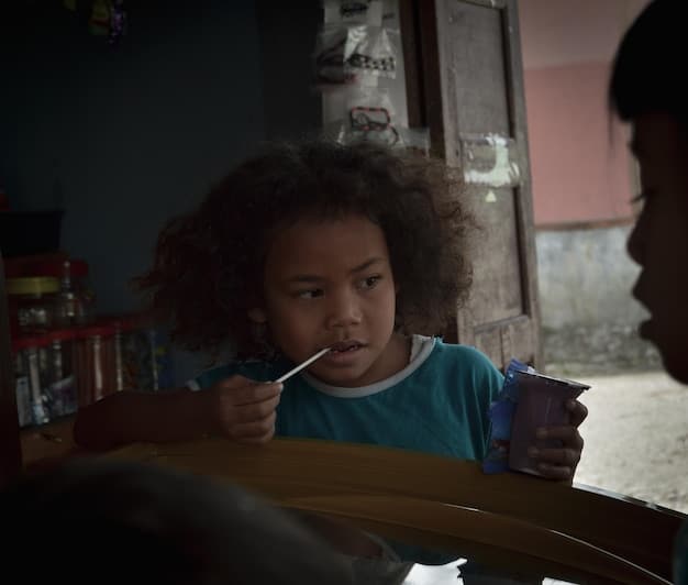 A close-up shot of a UN aid worker interacting with a child in a low-income neighborhood in the US. The aid worker is kneeling down, smiling, and offering the child a healthy snack. The background shows simple homes and a community garden, highlighting the local environment.